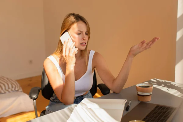 Tensed freelancer talking on smartphone near laptop and coffee at home in morning — Stock Photo