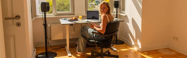 Blonde freelancer using smartphone near breakfast and laptop at home, banner — Stock Photo