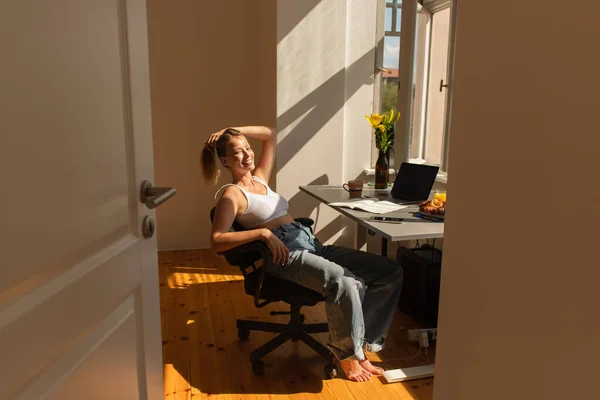 Happy barefoot freelancer looking at camera near devices and flowers at home — Stock Photo