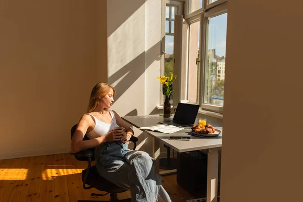 Side view of young woman holding coffee near devices and notebook at home — Stock Photo