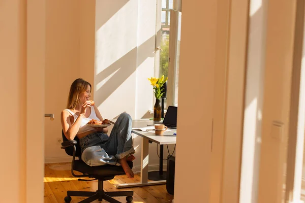 Freelancer positivo segurando croissant perto de laptop e café para ir em casa — Fotografia de Stock