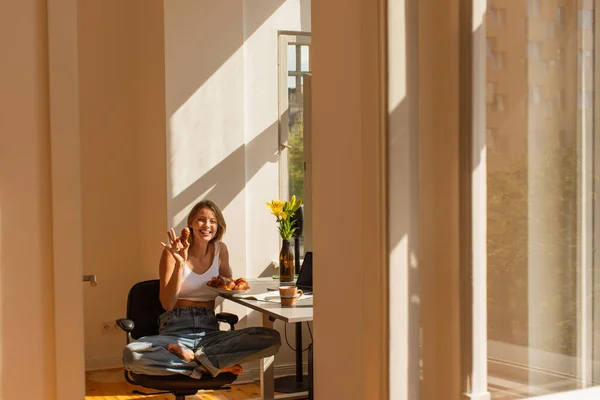 Donna sorridente che tiene il croissant e guarda la fotocamera vicino al computer portatile e ai fiori a casa — Foto stock