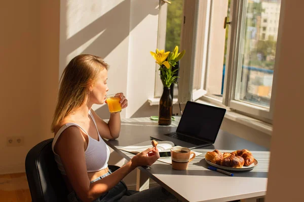 Vista lateral da loira freelancer beber suco de laranja perto de dispositivos e croissants saborosos em casa — Fotografia de Stock