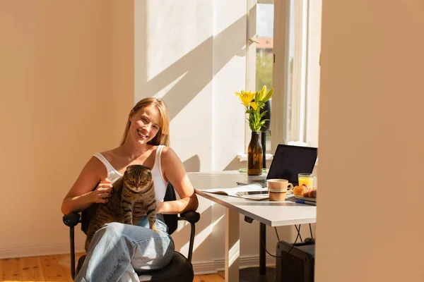 Lächelnde Frau hält schottische Faltkatze in der Hand und blickt in die Kamera neben Frühstück und Geräten zu Hause — Stockfoto