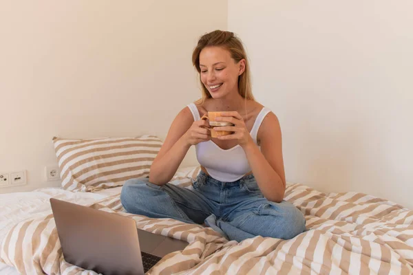 Mujer positiva sosteniendo taza de café cerca de la computadora portátil en la cama - foto de stock