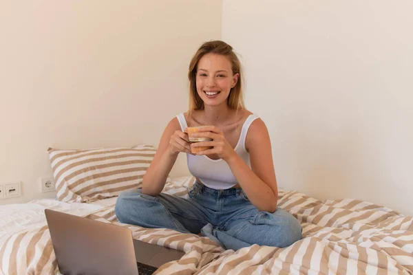 Веселая женщина в верхней части и джинсы, держа чашку кофе и глядя на камеру возле ноутбука на кровати дома — стоковое фото