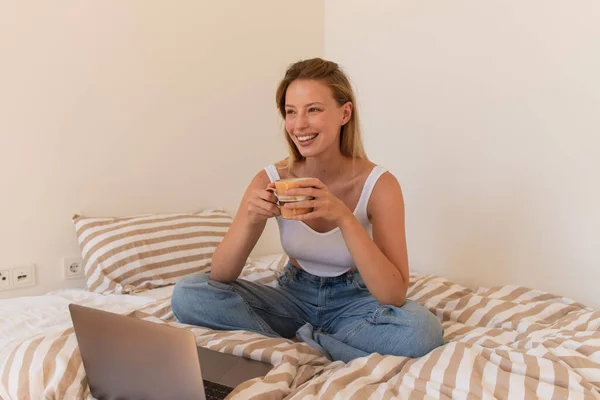 Mujer rubia feliz sosteniendo taza de café cerca de la computadora portátil en la cama - foto de stock