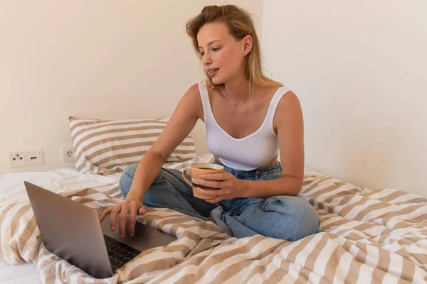 Blonde woman using laptop and holding cup of coffee in bedroom — Stock Photo