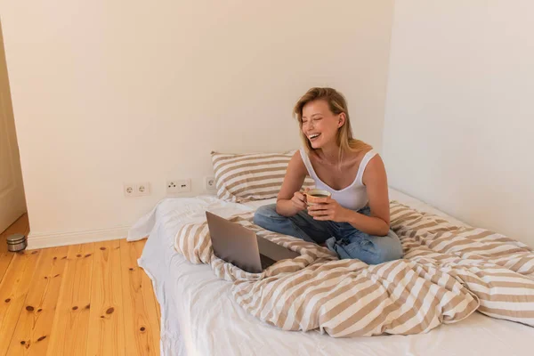 Весела жінка тримає чашку кави біля ноутбука на ліжку вдома — стокове фото