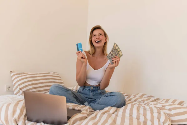 Jovem segurando cartão de crédito e dólares perto de laptop no quarto — Fotografia de Stock