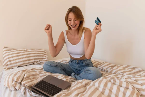 Femme excitée tenant carte de crédit près d'un ordinateur portable avec écran blanc sur le lit à la maison — Photo de stock
