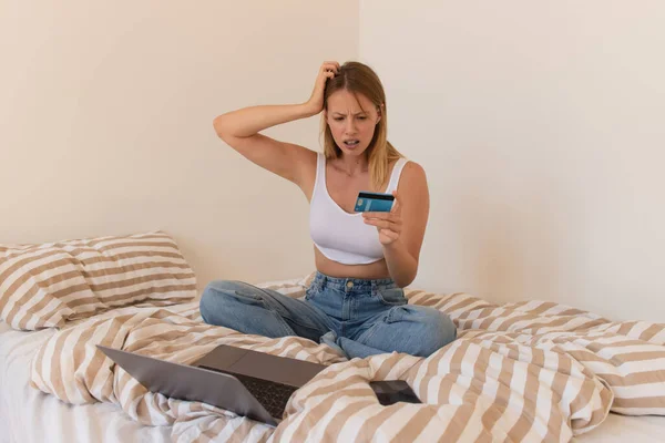Беспокойная женщина смотрит на кредитную карту рядом с устройствами на кровати дома — стоковое фото