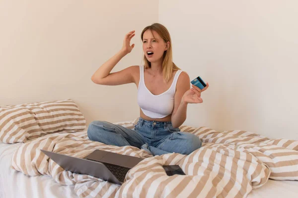 Недовольная женщина держит кредитную карту возле гаджетов на кровати дома — стоковое фото