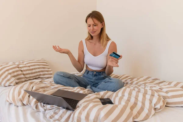 Расстроенная женщина держит кредитную карту возле ноутбука и смартфона на кровати дома — стоковое фото