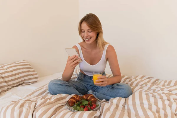 Smiling woman using smartphone and holding orange juice near breakfast on bed — Stock Photo