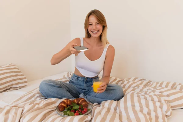 Mulher alegre segurando smartphone com tela em branco e suco de laranja perto do café da manhã na cama — Fotografia de Stock