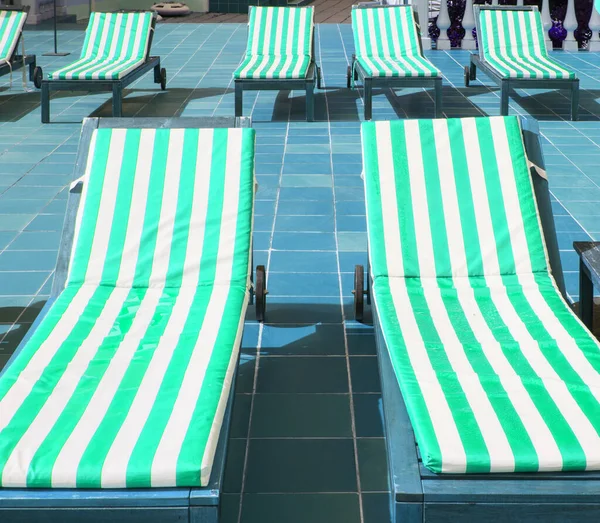 Empty wooden deck chairs with mattresses in green and white stripes stand in a row. Luxury outdoor recreation, sunshine.