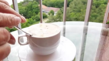 4K video, bir kadının eli sütlü bir fincan kahve kaldırıyor, yakın plan, kahvaltı, balkondaki cam bir masanın üzerinde.