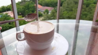 4K video, bir fincan sıcak kahve. Bir kadının eli lezzetli bir kapuçinoyu fincanda kaşıkla karıştırıyor, balkondaki cam masada içmek için yakın plan çekiyor..