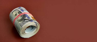 Many hundred dollar bills rolled up with a red rubber band on a brown background.