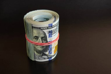 Many hundred dollar bills rolled up with a red rubber band on a black background.