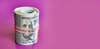 Many hundred dollar bills rolled up with a red rubber band on a pink background.