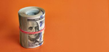 Many hundred dollar bills rolled up with a red rubber band on an orange background.