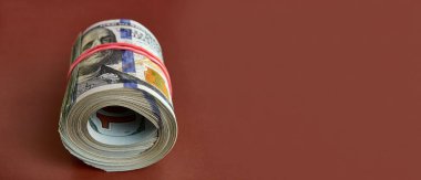 Many hundred dollar bills rolled up with a red rubber band on a brown background.