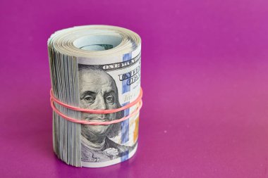 Many hundred dollar bills rolled up with a red rubber band on a pink background.