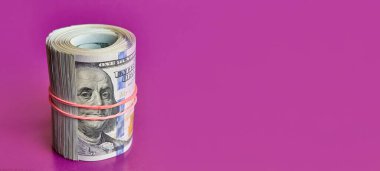 Many hundred dollar bills rolled up with a red rubber band on a pink background.