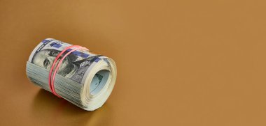 Many hundred dollar bills rolled up with a red rubber band on a gold background.