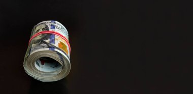 Many hundred dollar bills rolled up with a red rubber band on a black background.