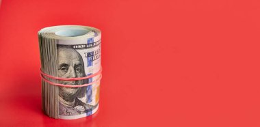 Many hundred dollar bills rolled up with a red rubber band on a red background.