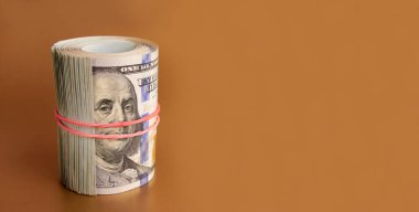 Many hundred dollar bills rolled up with a red rubber band on a gold background.