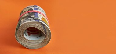 Many hundred dollar bills rolled up with a red rubber band on an orange background.
