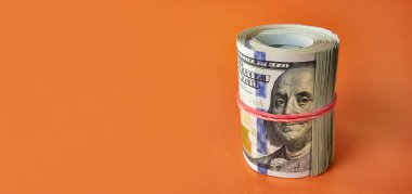 Many hundred dollar bills rolled up with a red rubber band on an orange background.
