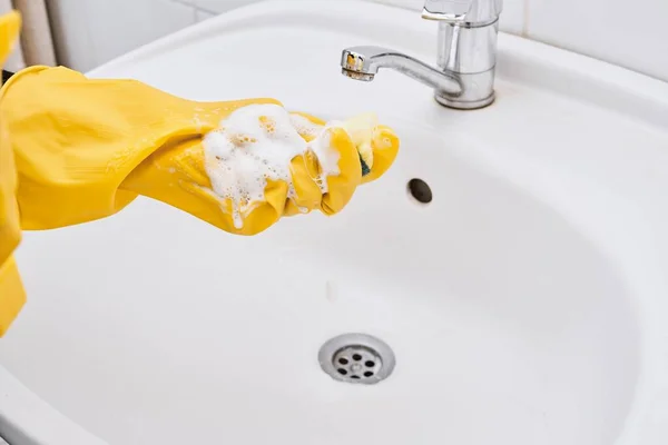 Cleaning a bathroom tap with sponge in yellow rubber gloves