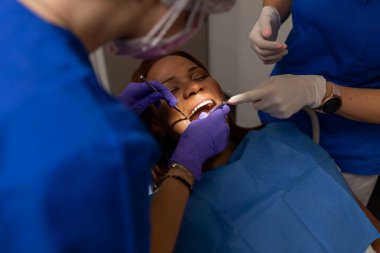 Siyahi bir kadın hasta ağzını açık tutarken diş hekimi diş eti ve diş etlerini kontrol ediyor ve diş eti hastalıklarını önlemek için diş eti ve diş etlerini temizliyor.