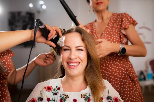 2人の美容師がヘアサロンでヘアドレッシングセッション中にヘアアイロンで丁寧にトリートメントしながら笑顔を見せる女性客 — ストック写真