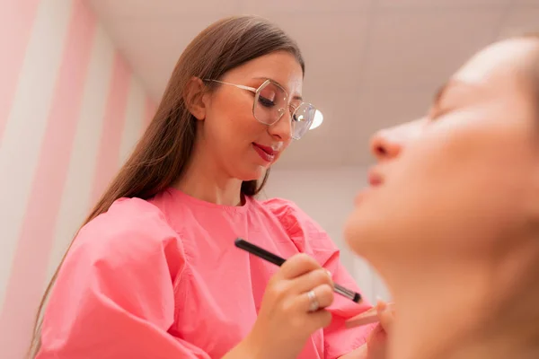 A beautician woman is brushing the foundation on the palette she is holding to apply it onto the customers face at the beauty salon