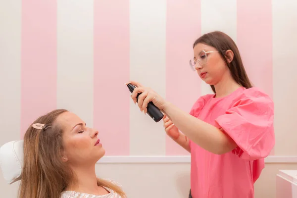 美容室で化粧セッション中にお客様の顔にファンデーションを吹きかける美容師の女性 — ストック写真