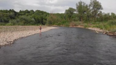 Sinema çekimleri post-processing olmadan (orijinal dosya, post prodüksiyon yok). Genç ve güzel bir kız nehirde duruyor ve bir oltayla balık yakalıyor. Açık bir günde nehrin berrak suları