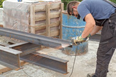 Elleri sıcak kıvılcımlardan korumak için koruyucu gözlüklü ve eldivenli bir adam inşaat alanında metal kesiyor.