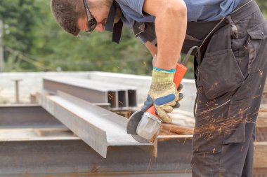 Elleri sıcak kıvılcımlardan korumak için koruyucu gözlüklü ve eldivenli bir adam inşaat alanında metal kesiyor.