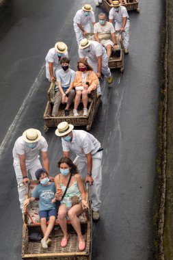 FUNCHAL, PORTUGAL - AUGUST 24, 2021: A group of unidentified tourists roll down the street in wicker baskets (toboggans) with the help of drivers in traditional uniforms.