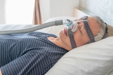 Orta yaşlı bir adam yatak odasında uyurken uyku apnesi için CPAP maskesi ve başlık takıyor.