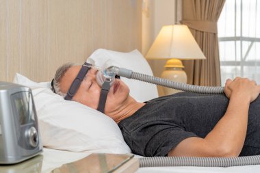 Orta yaşlı erkek, yatak odasında uyurken CPAP başlık takıyor ve maske takıyor.