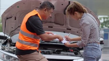 Worried young Asian woman talking to a car insurance agent or technician to claim the repair on her car accident and signing the claim form