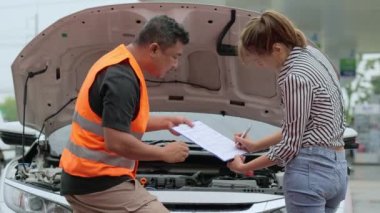 Worried young Asian woman talking to a car insurance agent or technician to claim the repair on her car accident and signing the claim form