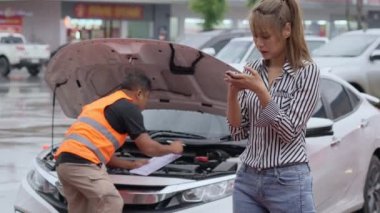Unhappy Asian female car owner standing in the rain at her car using her phone while the car insurance agent inspecting her car for the insurance claim report